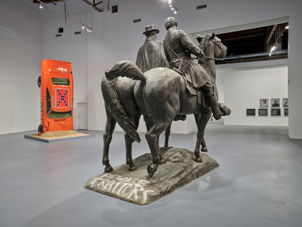 A large open gallery space with a large statue of two men on horseback in the foreground. In the background is a bright orange car with a Confederate flag painted on the roof standing on its hood as if it fell from a great height and crashed into the floor.