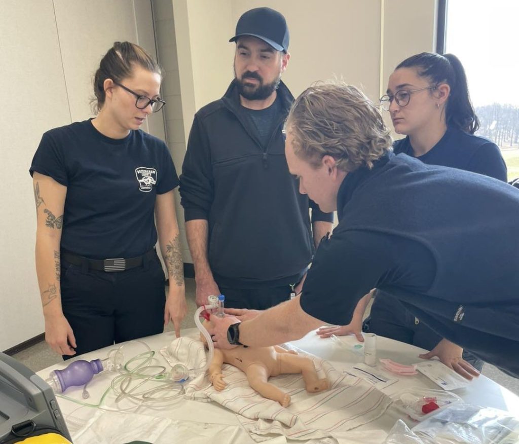 Two men and two women stand around a table as one of them holds an oxygen mask up to a small mannequin of a baby. Cloth and medical equipment litter the table