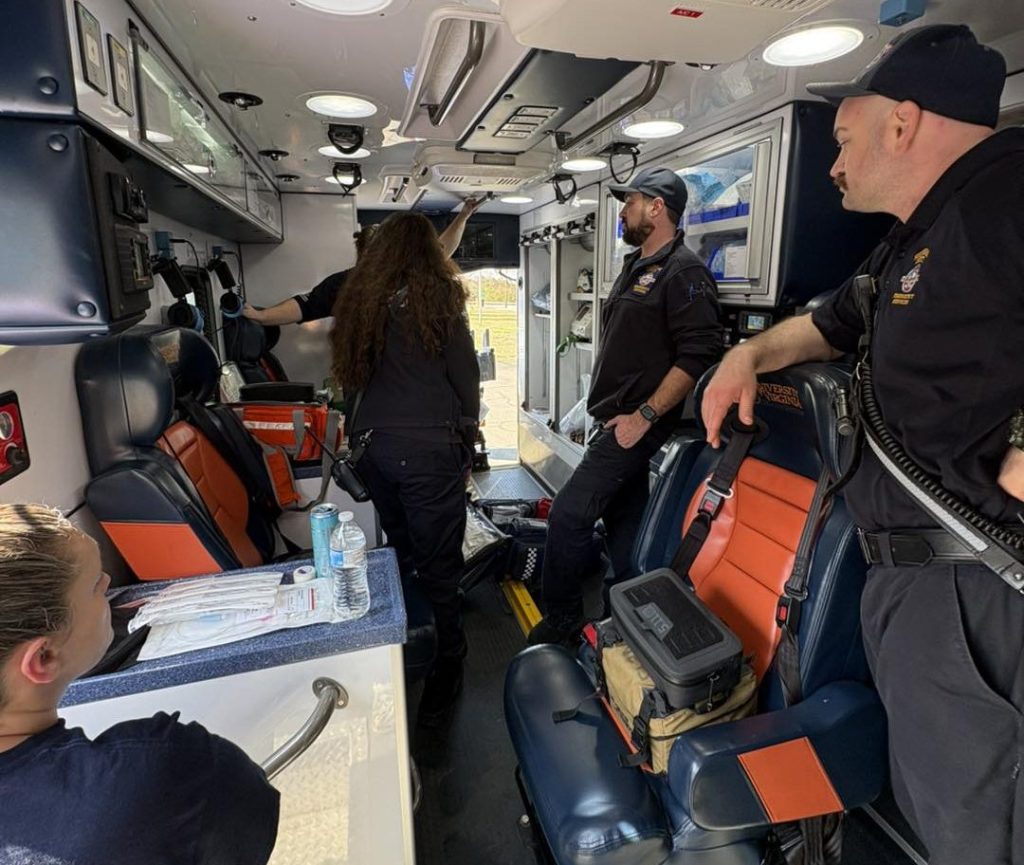 Five people stand in the back of a trailer filled with medical equipment, cabinets and captains chairs with shoulder-strap seatbelts.