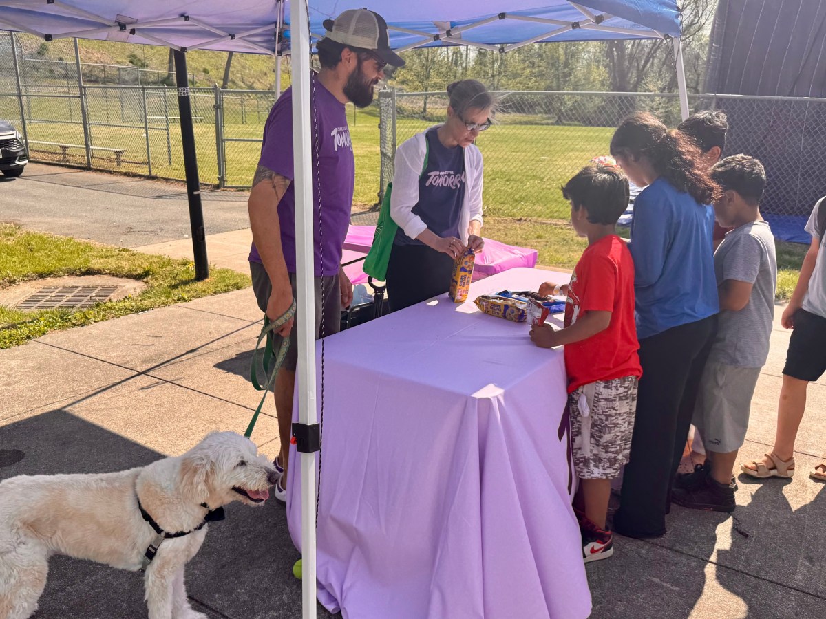 Two adults in matching t-shirts stand under a canopy tent. They talk to a group of children. In the bottom left, a fluffy dog. One of the adults holds the dog's leash.