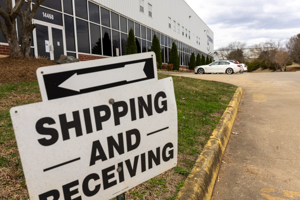 A black-and-white sign with the words 'shipping and receiving' and an arrow pointing left is in front of a light gray warehouse building with glass doors and windows.