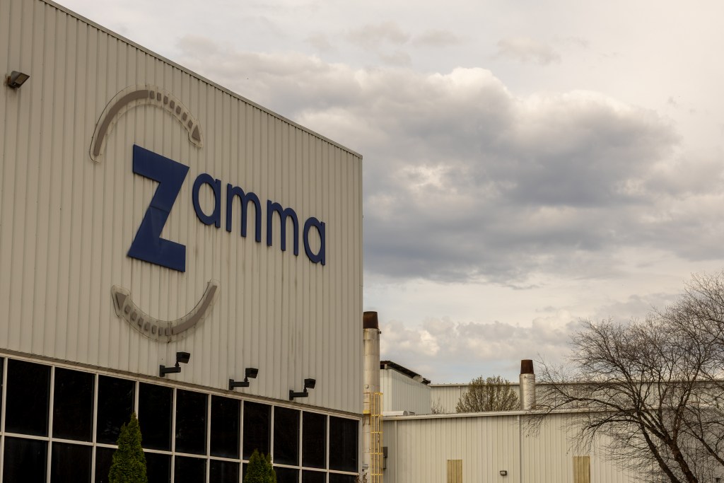 A white warehouse building with a large sign across the facade reading "Zamma." Windows line the ground floor with several security cameras visible above them.