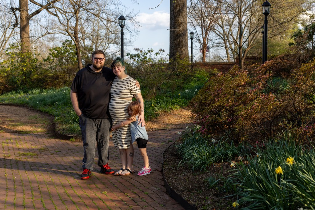A man in a black polo shirt stands on a brick pathway at a park with his wife, who is wearing a white-and-black striped dress, and their young daughter. Daffodils and other spring flowers can be seen in the background.