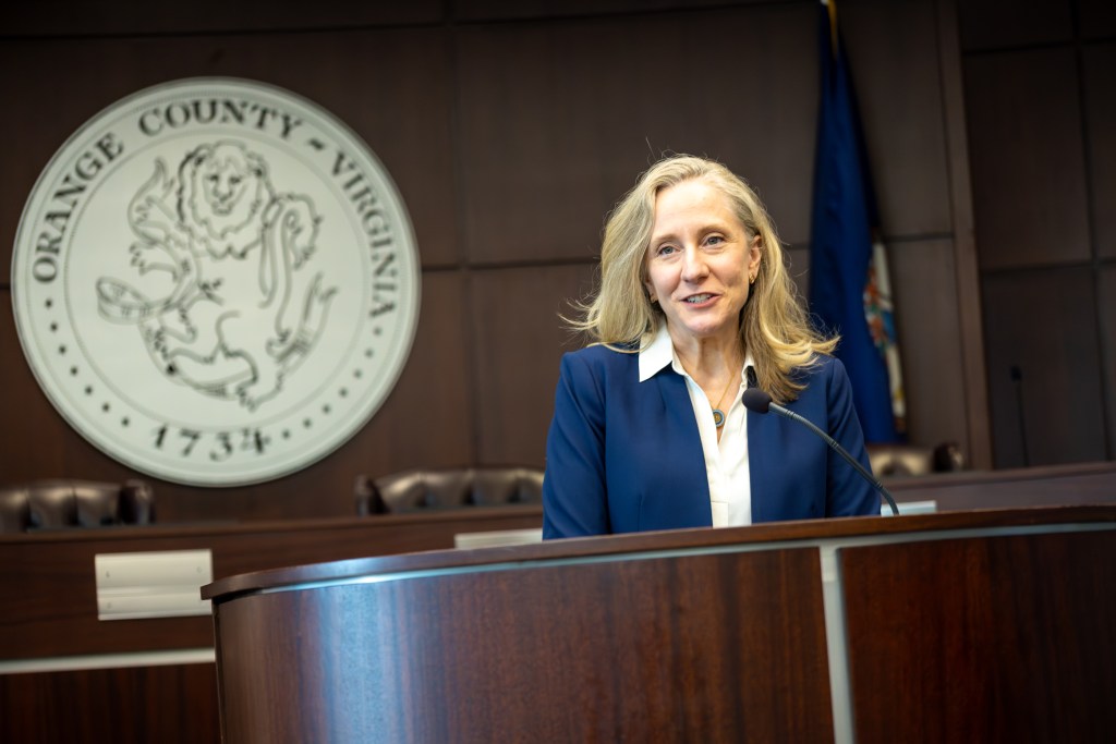 A woman with blonde hair and a dark blue suit speaks at a wooden podium. The official seal of Orange County, Virginia, featuring an illustration of a lion, is visible in the background.