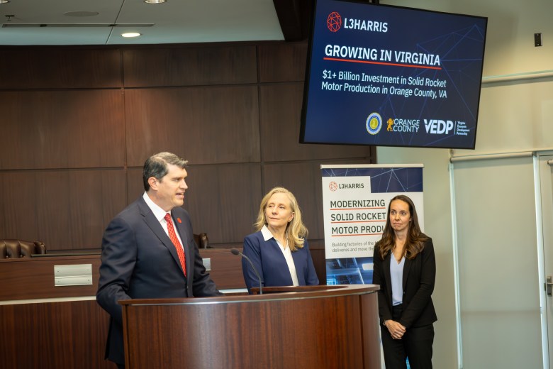 A man in a black suit and red tie speaks at a podium. At his right are the governor, who is wearing a navy suit, and another woman in a black suit. A television with the words "L3Harris: Growing in Virginia" is visible in the background.