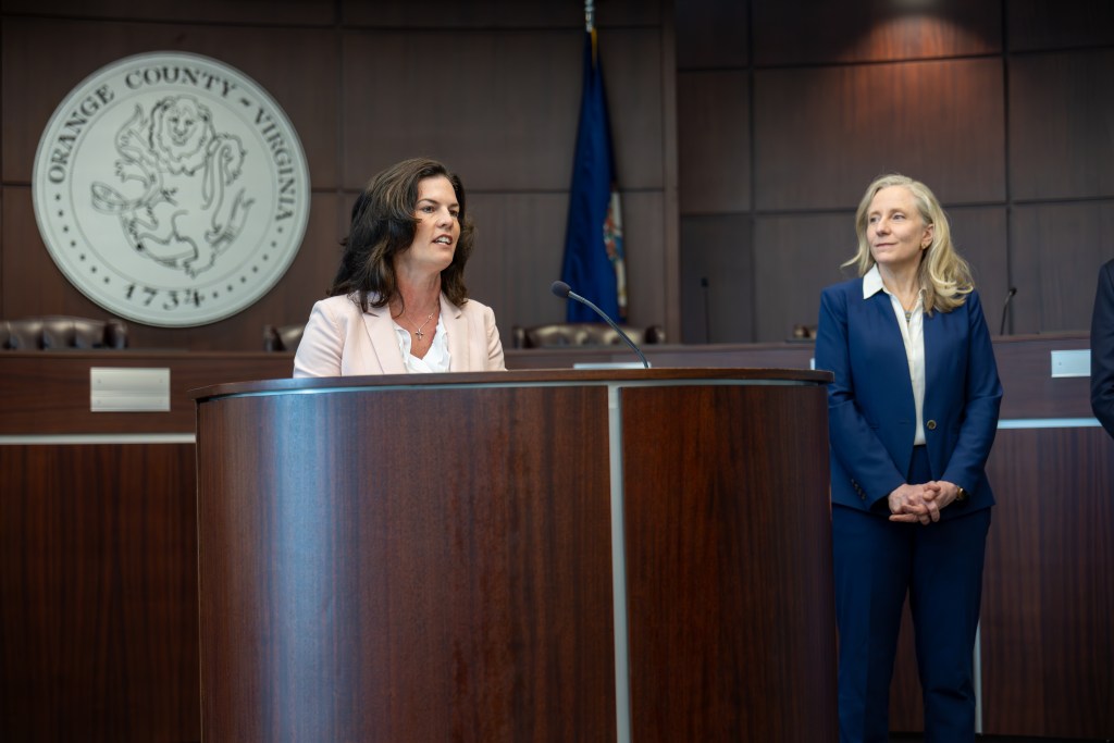 A woman in a pink blazer speaks at a podium. The governor, a woman in a dark blue suit, stands next to her. The official seal of Orange County, Virginia is visible in the background.
