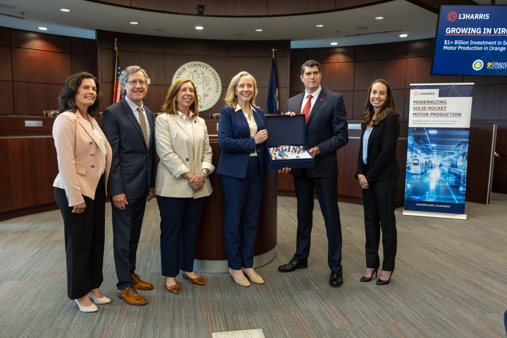 A woman in a blue suit and a man in a black suit hold a box containing a Virginia flag. Several other people in business attire stand beside them.