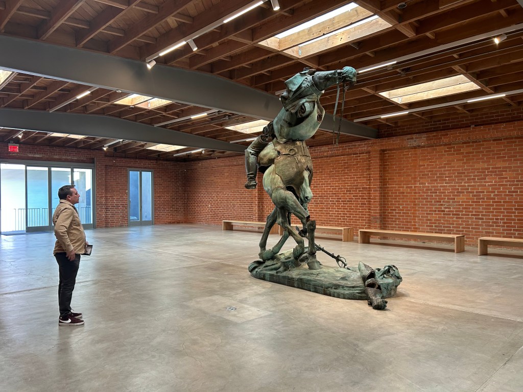 A man stands in a large open gallery space, looking at a statue that is made of parts of a soldier and a horse that are reassembled into a new uncanny creature.