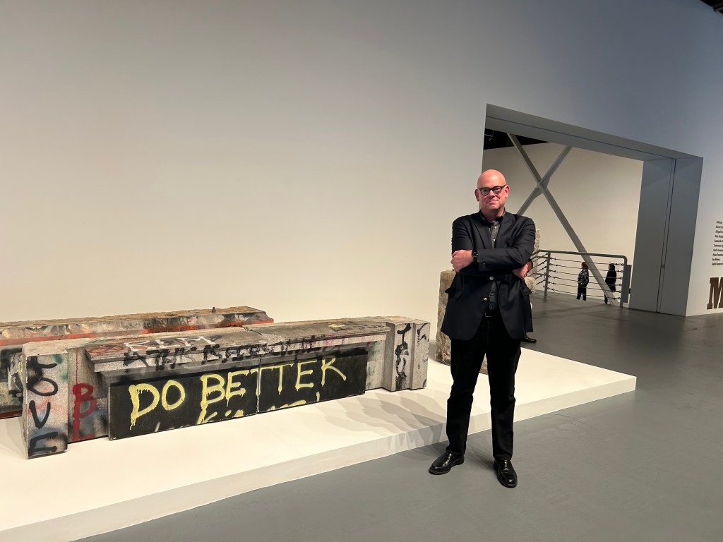 A man stands beside the hunks of granite, spray painted with the yellow letters, "Do Better." The slabs are on a white floor with museum lighting.