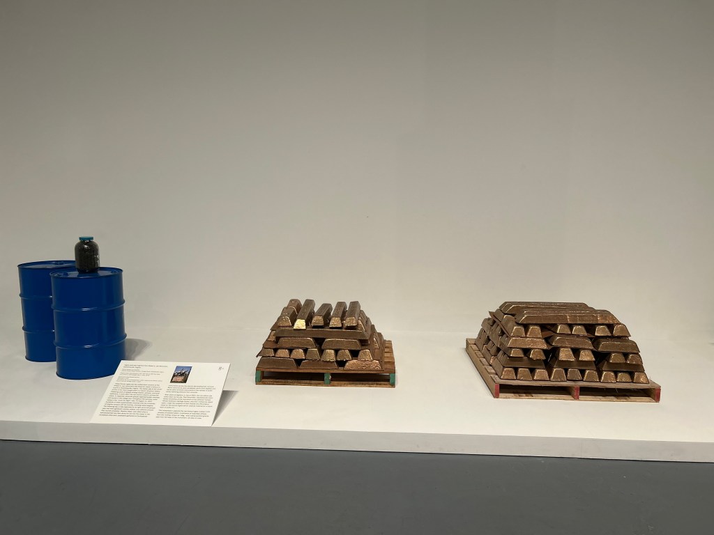 Bronze ingots are stacked in two piles resting on wooden pallets on the floor, with a clear glass jar of dark, dirt-like slag on a blue barrel beside them.