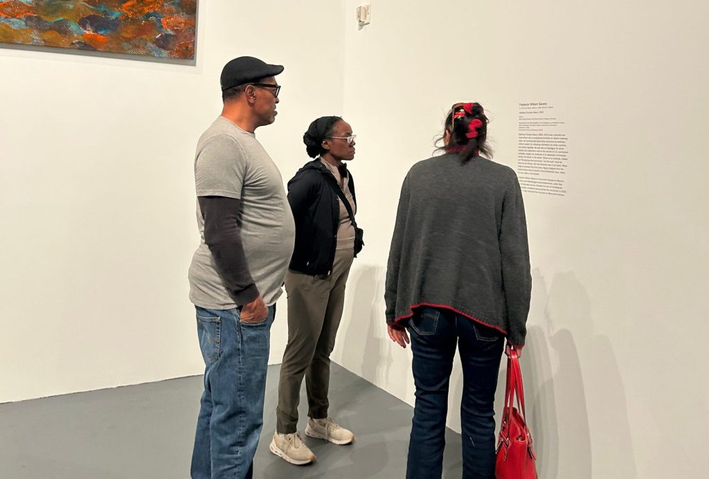 A man and two women stand in an open space in an art gallery, looking at a plaque with text that is hung on the wall near a corner of the room.