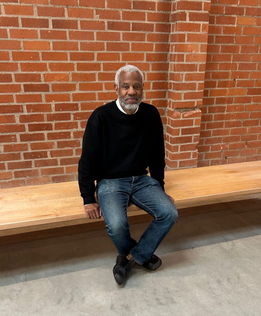 A man sits on a simple wooden bench looking at the camera. A brick wall is visible behind him.