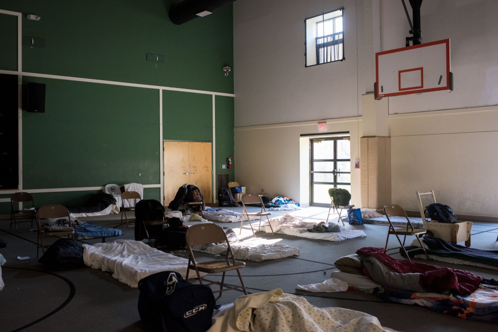 A photograph of a rec room being used as a temporary overnight homeless shelter. There is a dismantled basketball hoop hanging from the ceiling near the top right corner of the photo. On the floor of the room, there are cots and sleeping pads laid out on the floor. Some are covered with sheets and blankets, others with sleeping bags. Next to each one is a folding chair meant to serve as a spacer and bedside table.