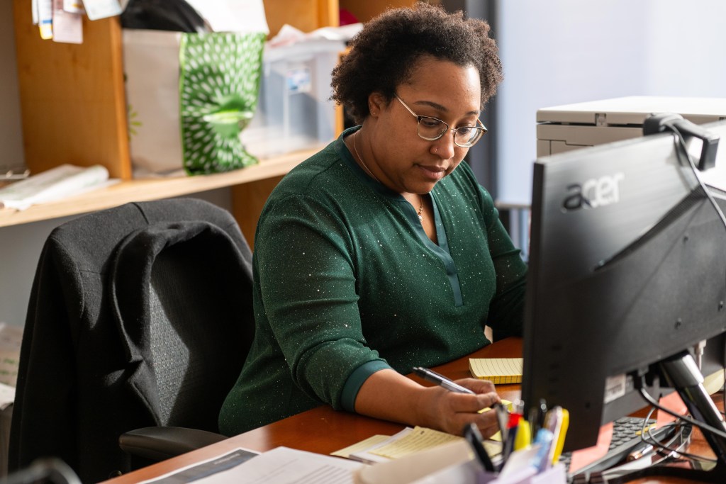 A photograph of a Black woman sitting at a desk in an office. There is a computer monitor in front of her, and she is focused on writing on a pad of paper.