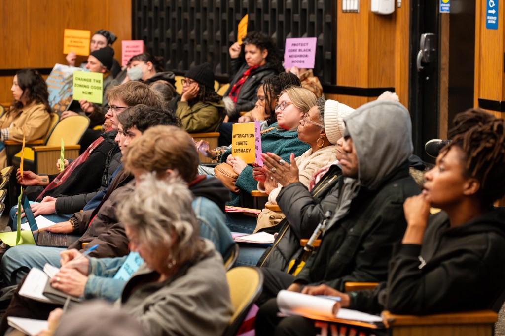 A photograph of people sitting in a public meeting. The group is diverse, featuring people of a variety of genders and races. Some of them hold up signs that say "Gentrification is white supremacy," "Protect core Black neighborhoods," "Change the zoning code now!" A few people are taking notes, and many of them are listening with exasperated expressions.