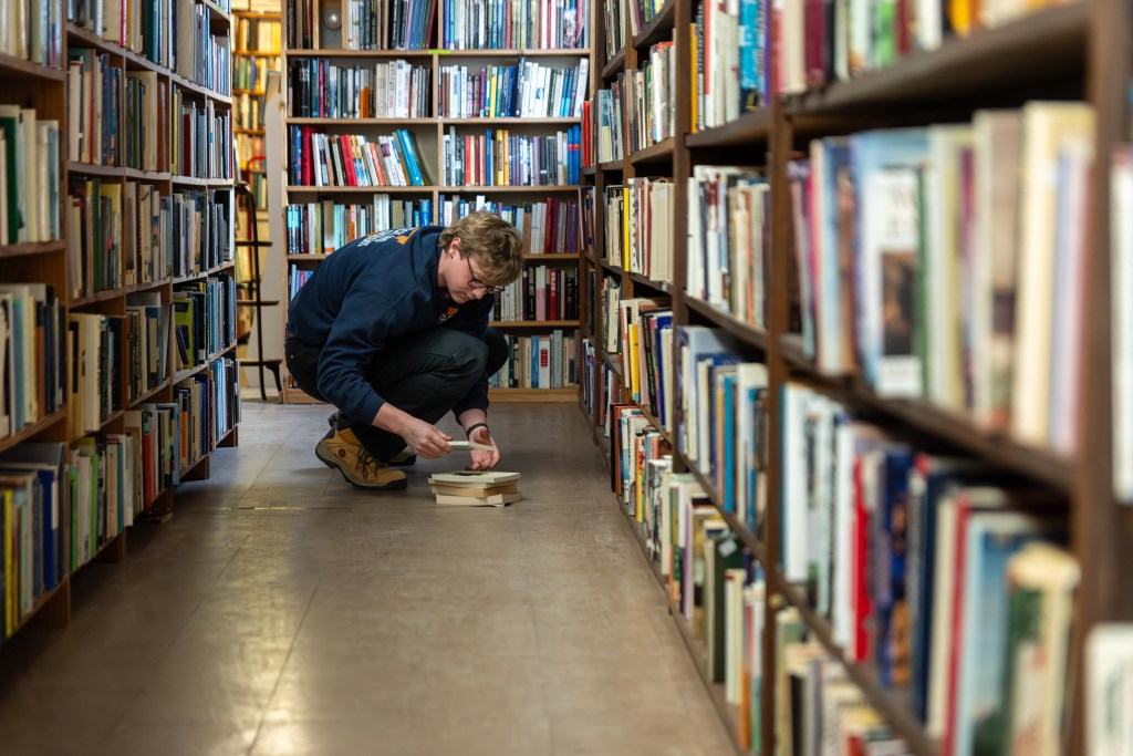 A young man in a navy UVA sweatshirt leans over a small pile of books on the floor. Behind him and on either side of him are wooden shelves filled with books.