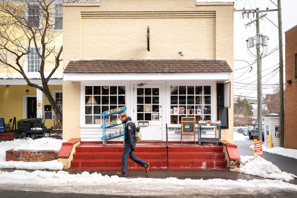 A black chalkboard easel that reads "Ginkgo Books" and two rolling utility carts full of books are positioned outside a yellow brick building with a white facade, brown slated roof and bright red stairs. A man wearing a backpack walks by.