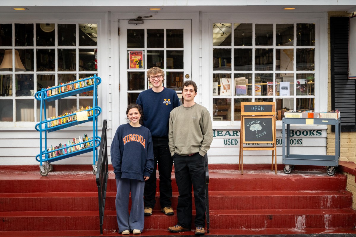 One young woman and two young men stand on red stairs leading up to a yellow brick building with a white facade and brown slated roof. Beside them are two rolling utility carts full of books, and an easel that reads "Ginkgo Books."