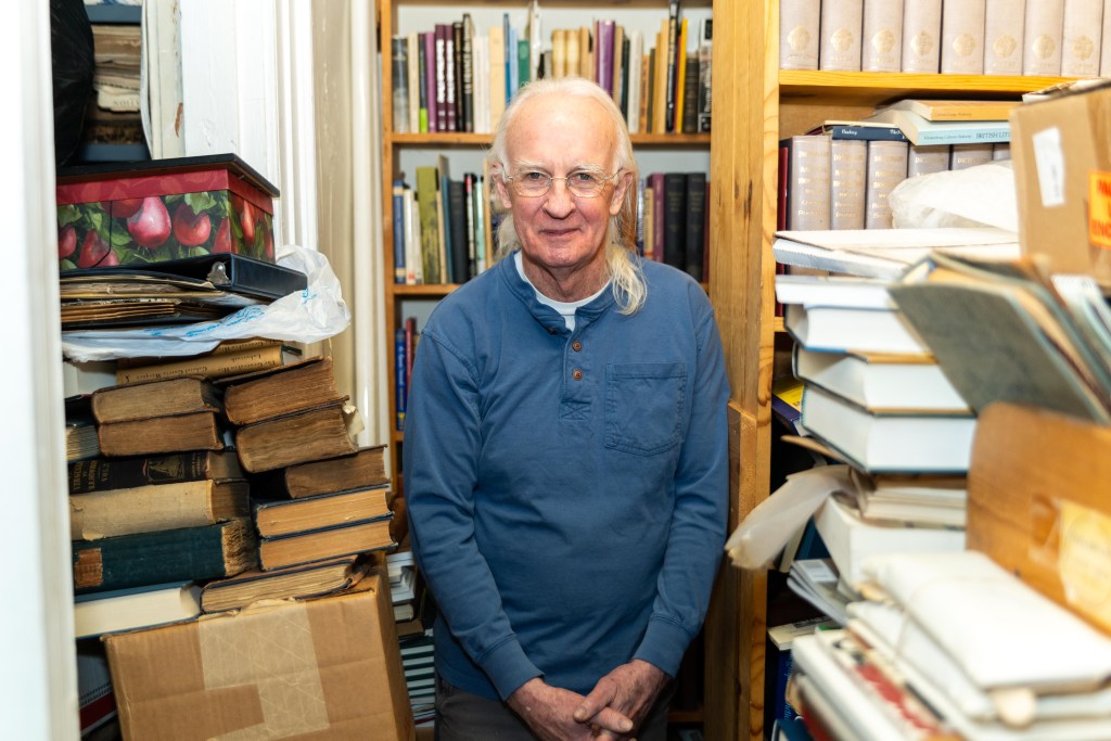 A man with long white hair, wearing glasses and a long-sleeved blue shirt, stands in front of and beside two yellow-brown wood shelves filled with books. He is surrounded by stacks of books.