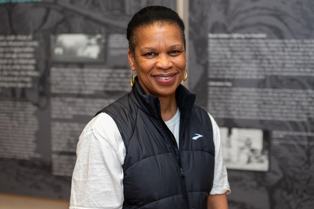 A woman wearing a white short-sleeved shirt and black vest looks at the camera, smiling. Behind her, large signs with text and photos are visible but out of focus.