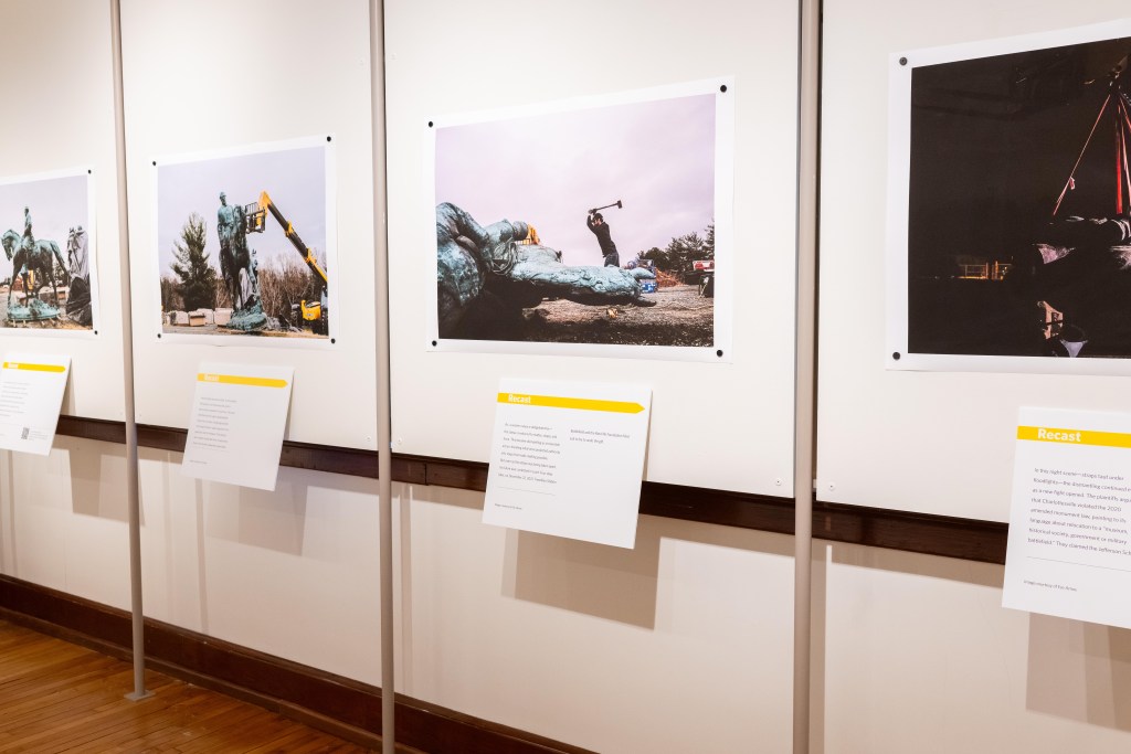 A series of large photos and descriptive text hung on a wall, depicting a large statue being taken down and broken apart.