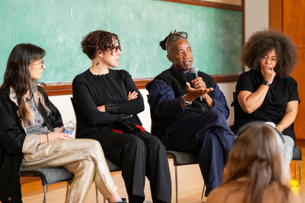 Three women and a man are seated in chairs organized in a circular shape. The man is holding a microphone and speaking while gesturing with his free hand.