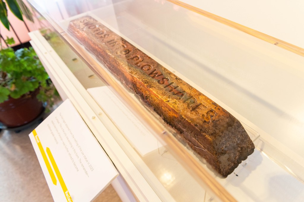 An aged and dirty block of wood with with the words "swords into plowshares" carved into it sits in a display case in a brightly lit room.