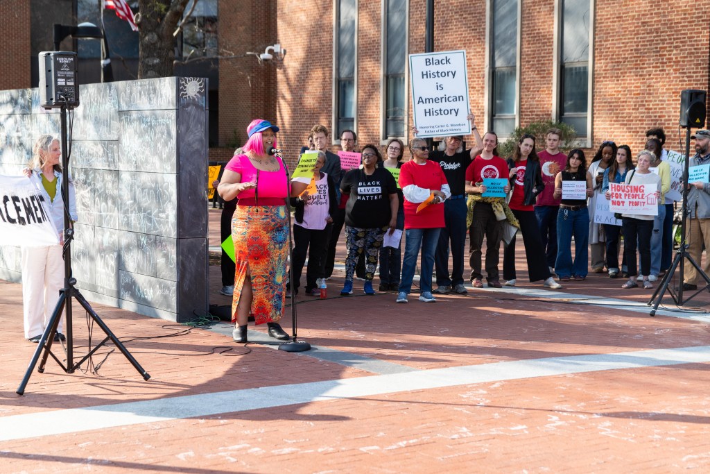 A photograph of people gathered outside for a rally. They're on a brick pedestrian mall, standing in front of a brick municipal building and a "free speech" wall covered in chalk messages. At the center of the photo, a woman wearing brightly-colored clothing speaks into a microphone while holding a sign that says "Change the Zoning Now!" Other signs say "Black History is American History," "Housing for People, Not Profit," "No LV Collective No Mark," and "No Luxury Apartments While People Are Unhoused." Two people hold up a long cloth banner that reads "No Displacement."