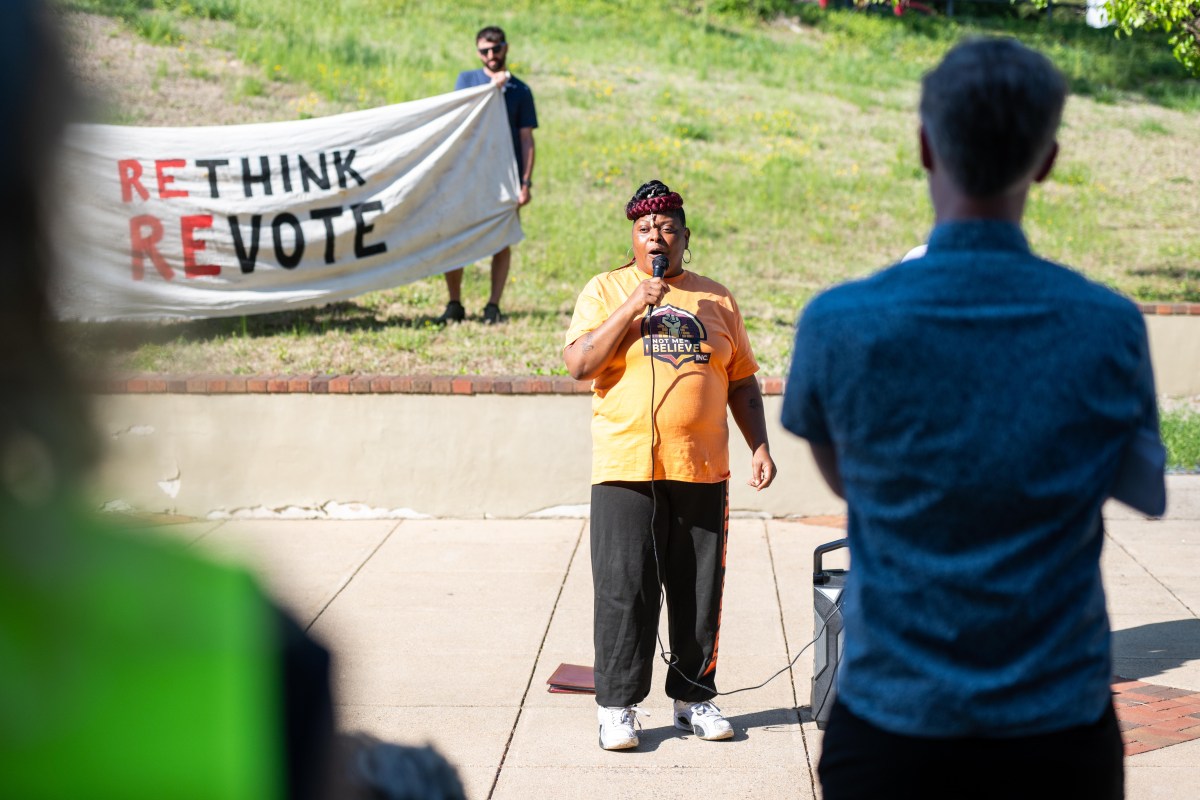 A woman stands outside speaking into a microphone as people in the audience watch and listen. Two people are standing behind her holding a banner that reads "Rethink, Revote."