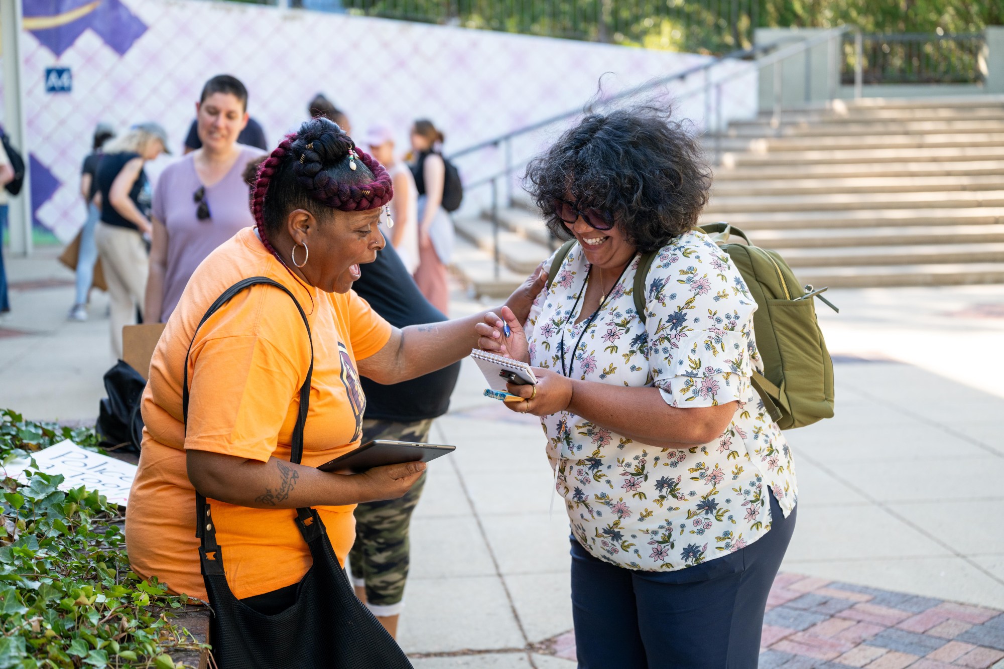 A woman in a t-shirt and large hoop earrings speaks with another woman, touching her arm. The other woman smiles and takes notes on a notepad.