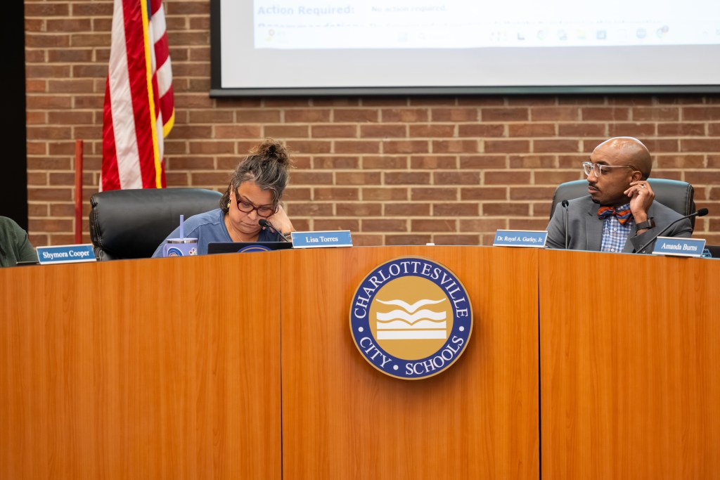A woman sitting at a dais looks down while leaning her head against her hand. A man sits to her left and looks at her.