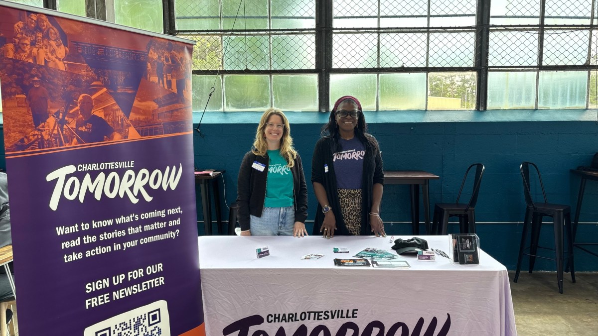 Two women stand behind a table. They wear t-shirts with the same logo that reads TOMORROW. On the left of the image is a banner with the logo CHARLOTTESVILLE TOMORROW.