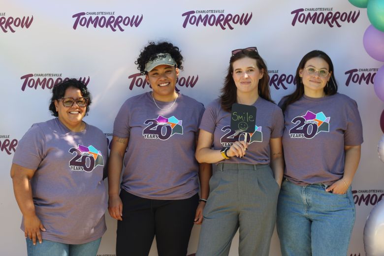 Four people smile at the camera. They were matching shirts that read: NEXT 20. They stand in front of a banner with repeated CHARLOTTESVILLE TOMORROW logos. There are balloons to their left and right.