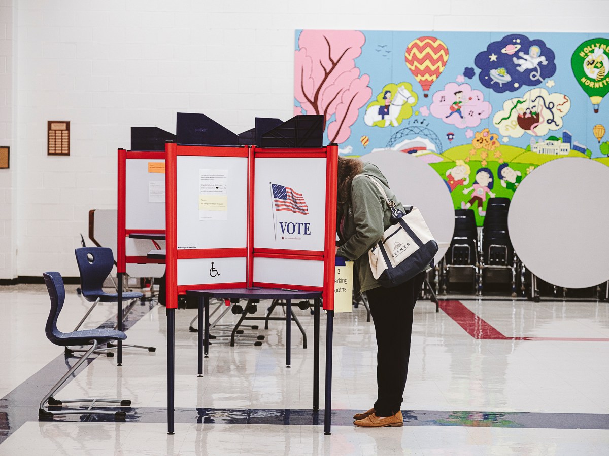 Tables and chairs are set up in a large open space with privacy dividers featuring an American flag and the word "vote." A woman with a canvas bag stands a table leaning forward so her face is hidden behind the divider.