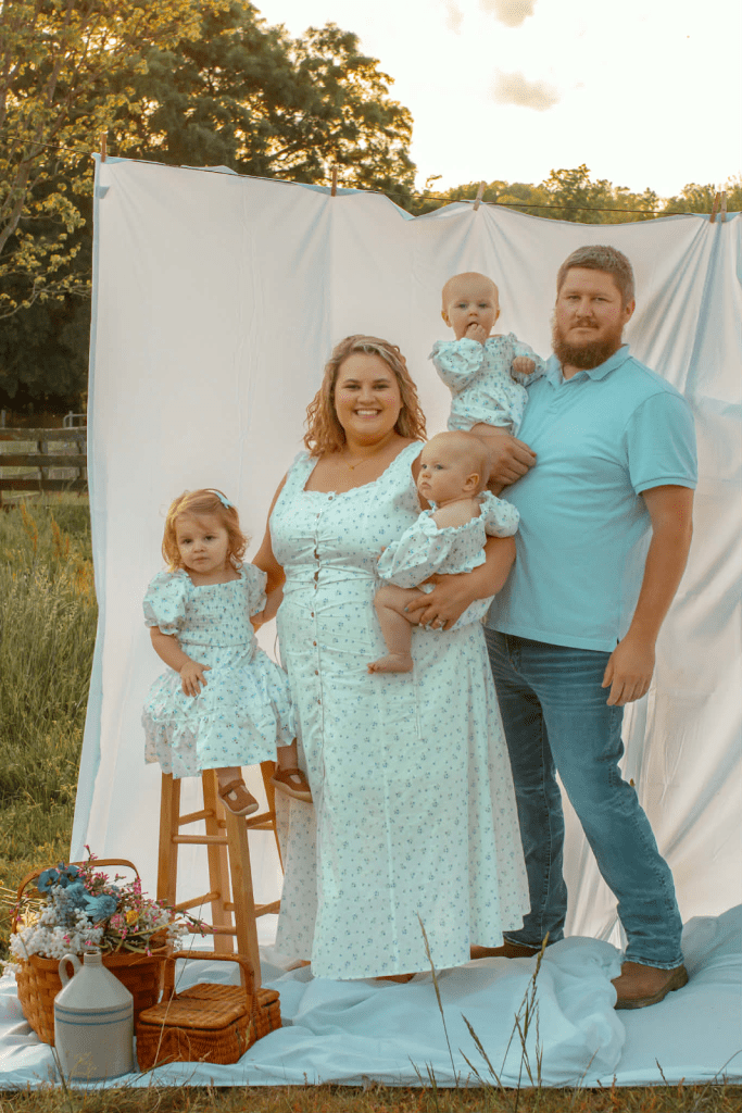 A family is posing in a field in front of a white sheet. The woman is holding two kids — an older girl and a twin. Her husband next to her is holding the other twin. The woman and the kids are wearing matching outfits.