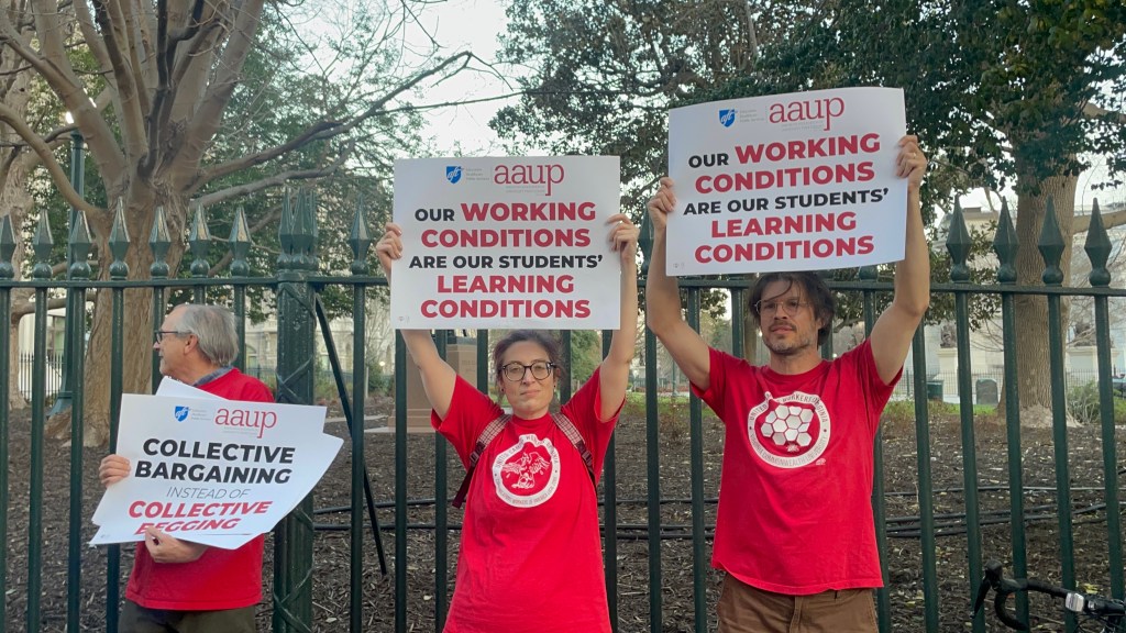Members of Virginia higher-education unions, including some from the University of Virginia, rally at the Virginia Capitol on March 11, 2026, for full inclusion in collective bargaining legislation that they say is necessary to enable them to negotiate for better pay, to keep up with rising costs of living, and better working conditions. ALT: Three people in red shirts stand in front of a black iron fence and hold up signs that read "our working conditions are our student's learning conditions" and "collective bargaining instead of collective begging."