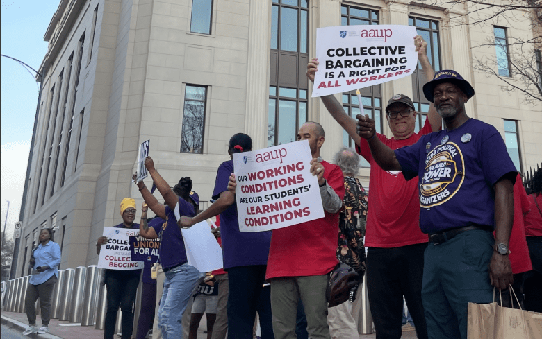 A line of people in red and purple shirts hold up signs expressing support for collective bargaining and improved working conditions.