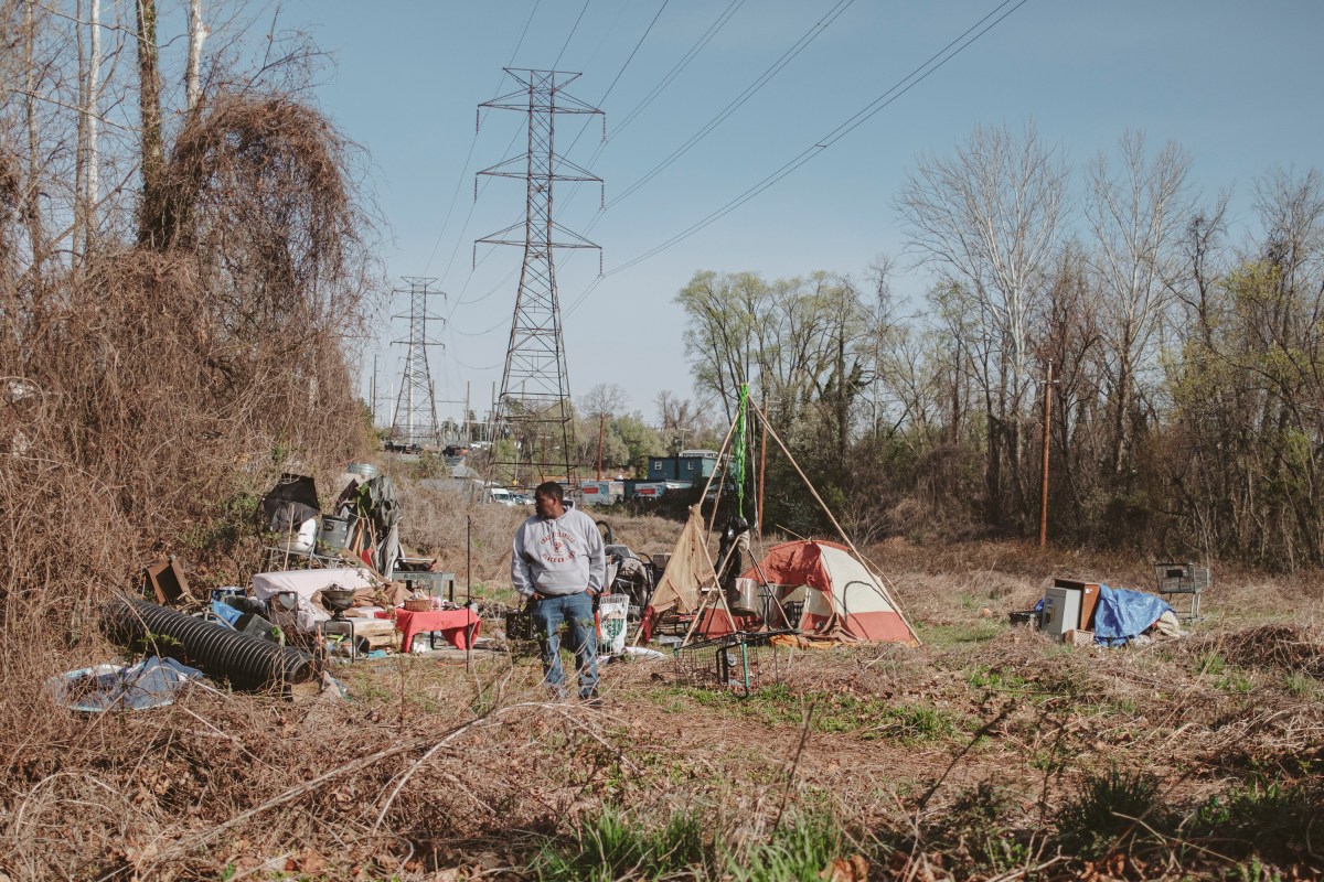 A man stands at a homeless encampment with his hands in his pockets and his gaze cast to one side. The landscape around him is mostly brown and overgrown, with a few trees budding in the background. There is a tent nearby, surrounded by piles of things, including a few grocery carts, small tables, a woven basket, and what appears to be a futon covered by a bedsheet.Transmission towers are visible in the background and power lines run overhead.