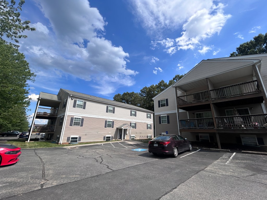 Two three-story residential buildings with wooden patios and two cars in a parking lot, with trees and a bright blue sky behind them.