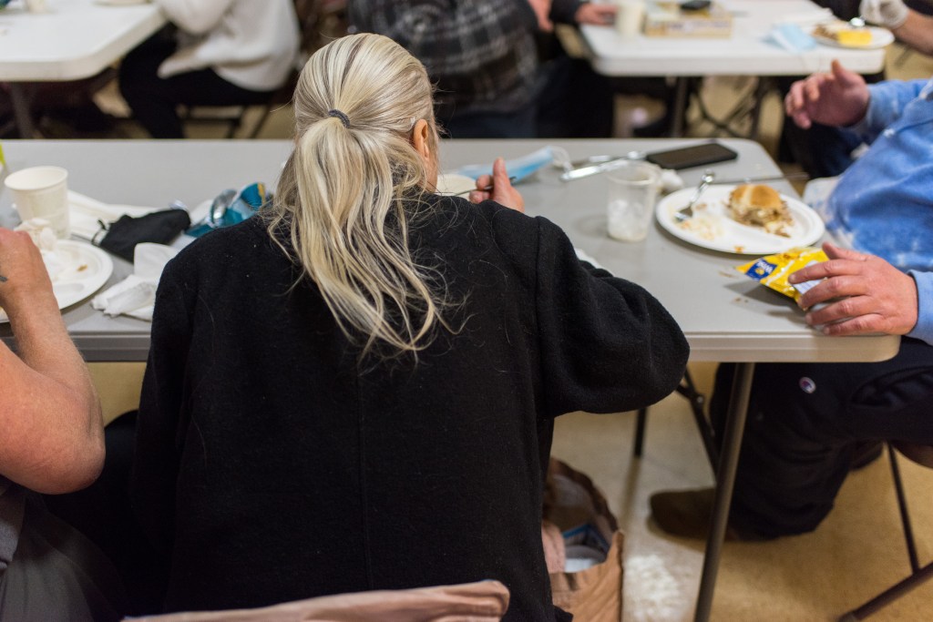 A photograph of people sitting around folding tables in a church recreation area. At the center of the photo is a person eating dinner from a paper plate. They're wearing a fleece jacket, their hair pulled back into a low ponytail. The arms and hands of other people are also visible, but out of focus.