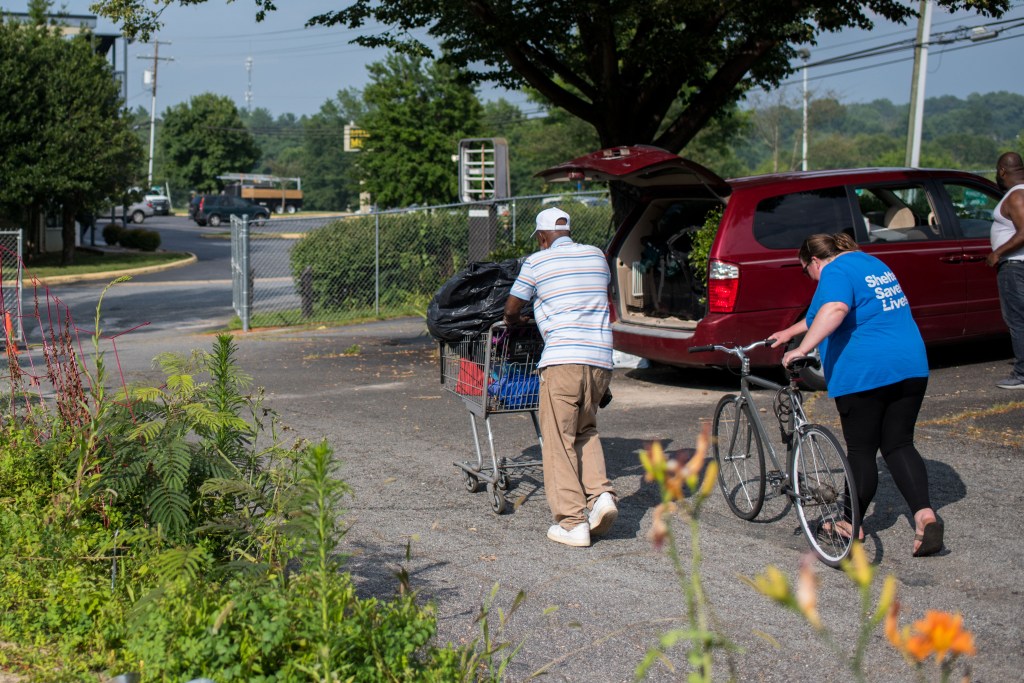 A photograph of two people, an older Black man and a middle-aged white woman, pushing a shopping cart and a bicycle across a parking lot. Neither person's face is visible. The man wears a baseball cap, a polo short, khaki pants and white sneakers. He pushes the shopping cart, which holds a few small items and, on top of them, a large black trash bag full of his belongings. The woman walks behind him, pushing a bicycle. She wears sandals and leggings, and a t-shirt with "Shelter Saves Lives" printed on the back. Another person loads a minivan nearby. The trees and plants around them are green, and a highway is visible in the background.
