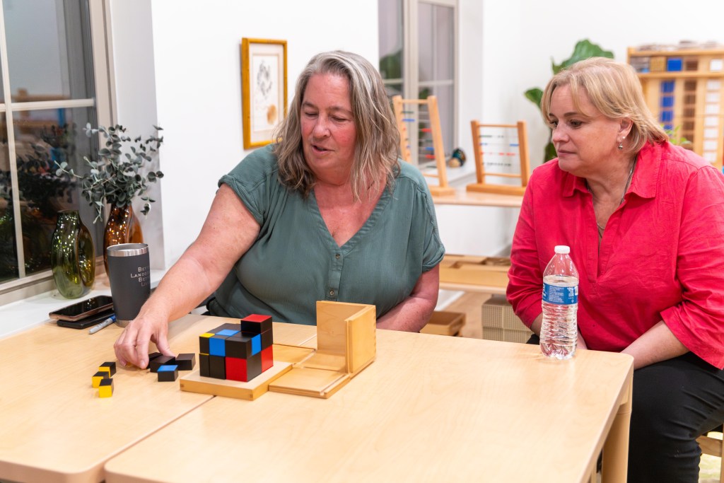 A woman shows how to use a trinomial cube, a colorful set of blocks, as another looks on. Both sitting in wooden children's furniture.