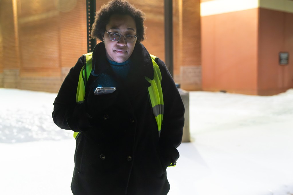 A Black woman stands outside a grocery store loading area. She wears glasses, a winter coat, gloves, and reflective safety vest. It's nighttime, and bright lights from the brick building behind her illuminate the sheet of snow and ice covering the parking lot. She is having a conversation with someone on her phone, via speakerphone, and has a sad look on her face.