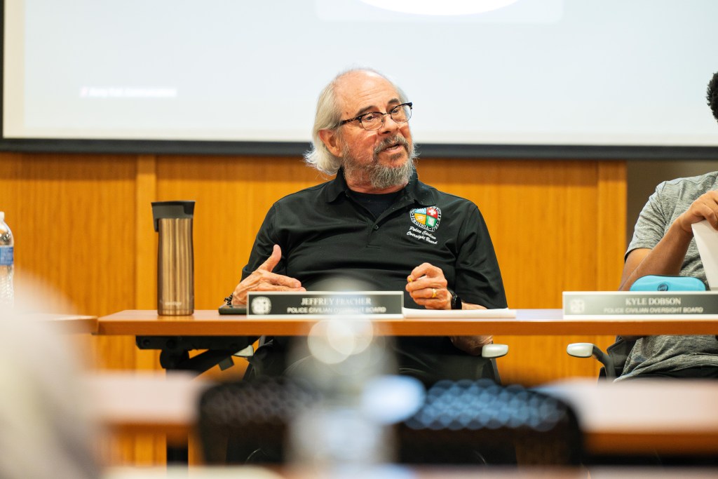 A man with glasses and a beard wearing a black polo shirt sits at a table and looks to his left. He is speaking and gesturing with his hands.