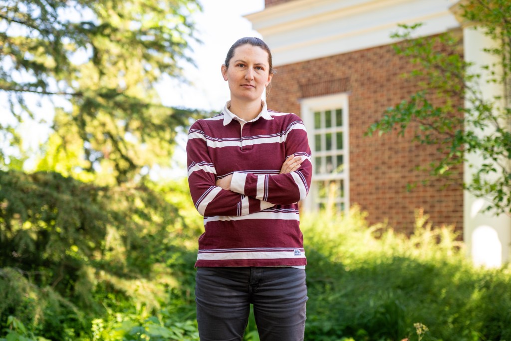 A woman with a striped red and white collared shirt stands with her arms crossed in front of green foliage and a partially obscured red brick building with white trim.