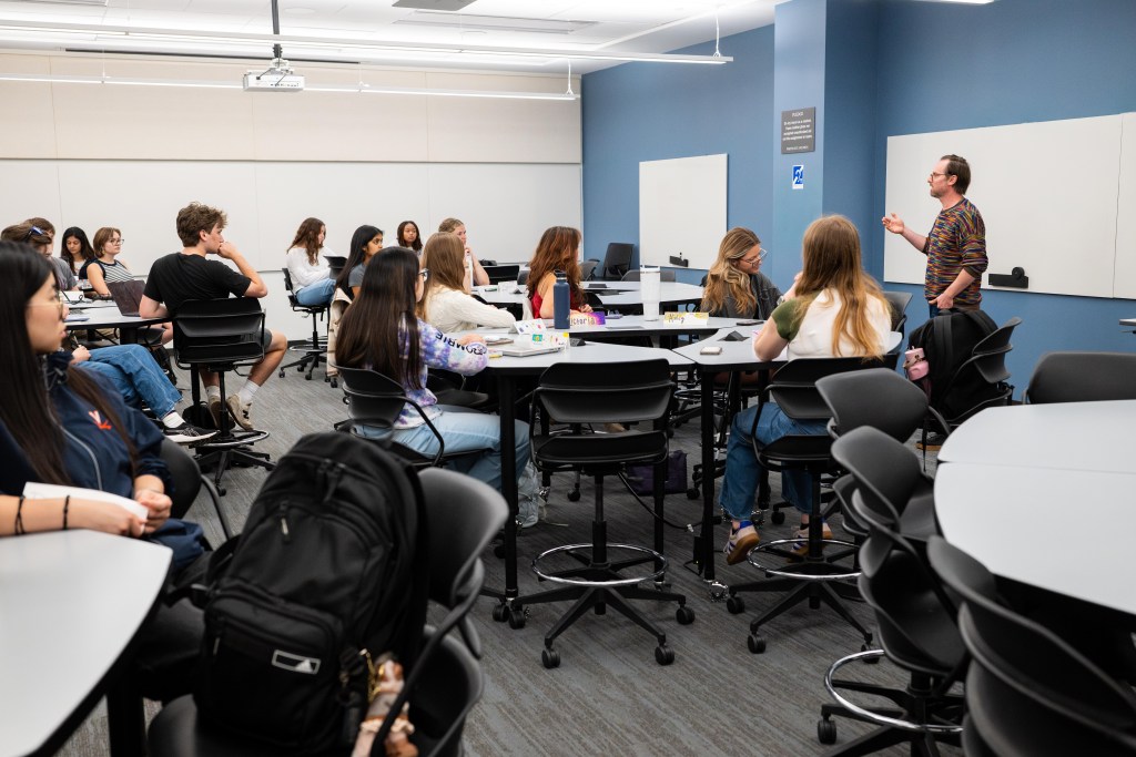 Several young adults sit in groups across five tables in a classroom with grey carpeting and alternating white and light blue walls, facing a man standing in front of a whiteboard and gesturing toward them with one hand.