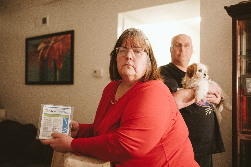 A woman in a red blouse holds up a laminated flier that reads "Adviron Cockroach." A man holding a small dog stands behind her. They are in the living area of a home, with artwork on the wall behind them above a sofa.