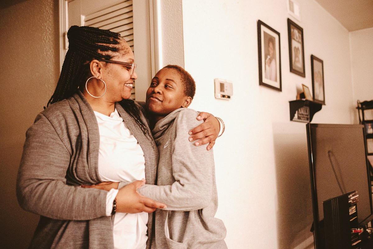A woman in hoop earrings and a brown sweater embraces a young woman who has a gray sweatshirt. They are in the living area of a home with pictures on the wall and a television off to the side of the image.