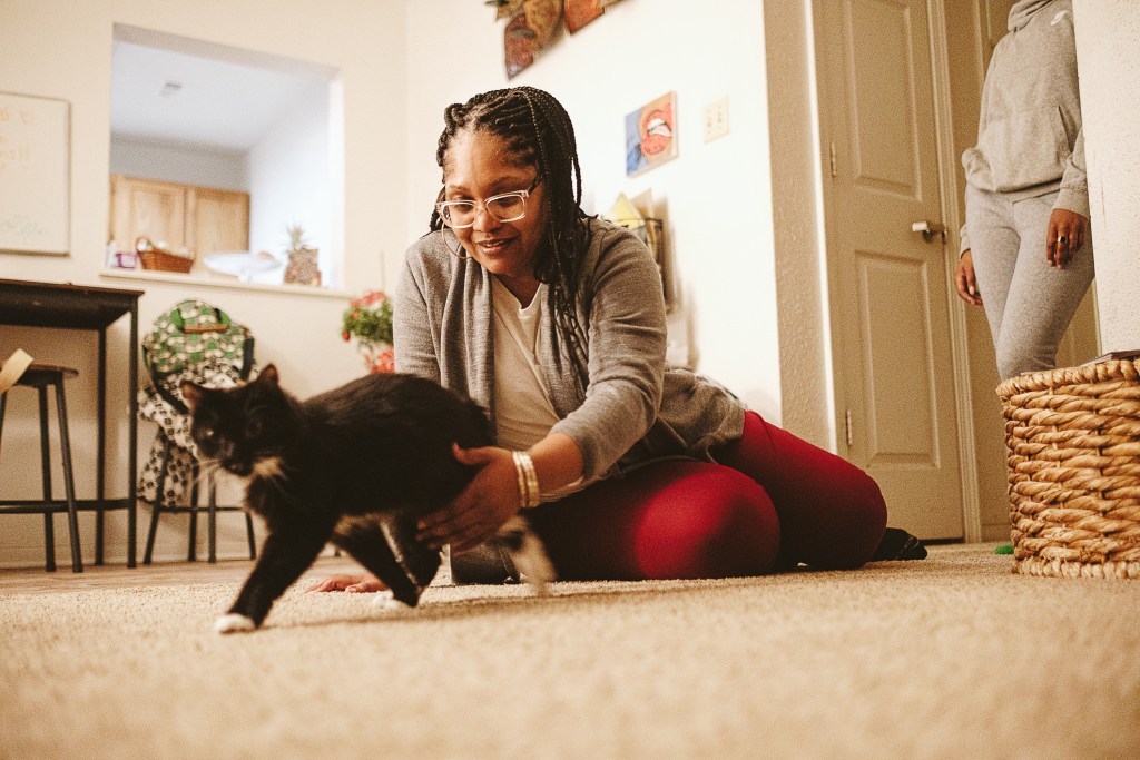 A woman sitting on a brown carpet in a living room reaches to hold a black and white cat that is looking at the camera. In the background are stools and a table in front of a window to another room, and a person in a grey sweatsuit off to the side.