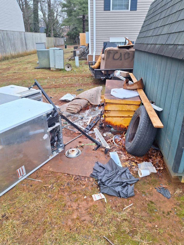 A pile of old appliances, a tire, pieces of wood, a rug, trash piled up next to a shed, with. building behind.